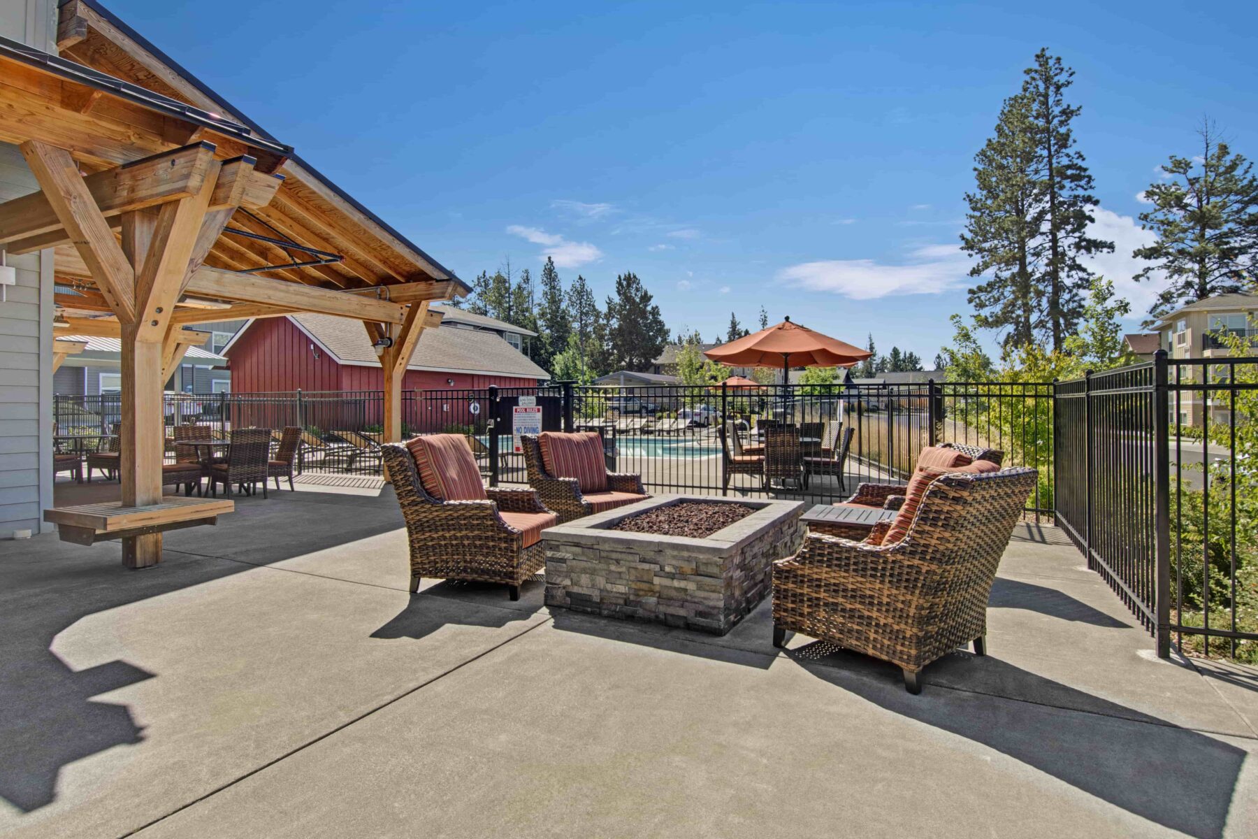Outdoor patio with fire pit, wicker chairs, wooden pergola, red umbrella, picnic table, and red barn-like building in background.