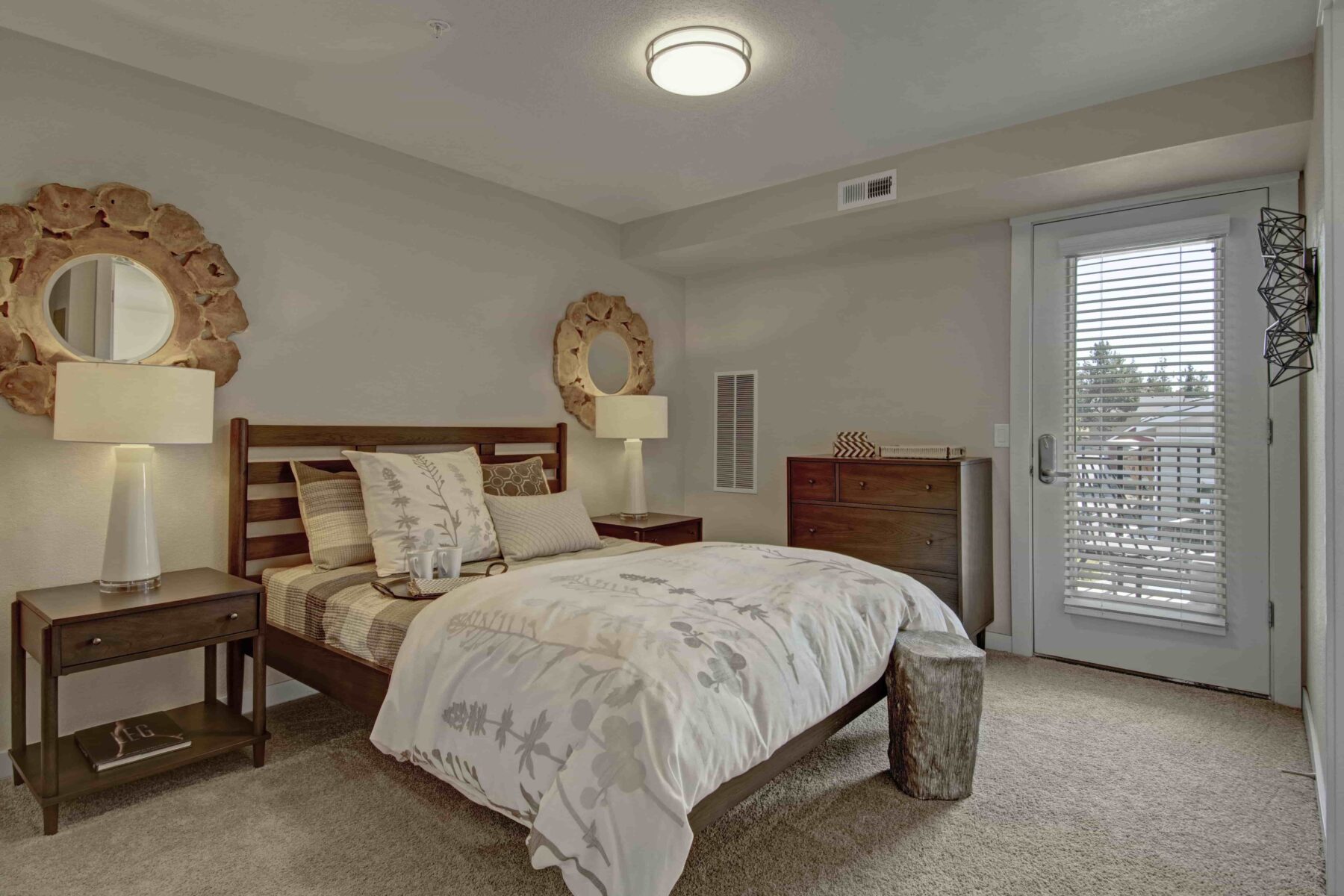 Bedroom with queen size bed, wooden bedside table, white lamp, round mirror, and white/gray furry rug.