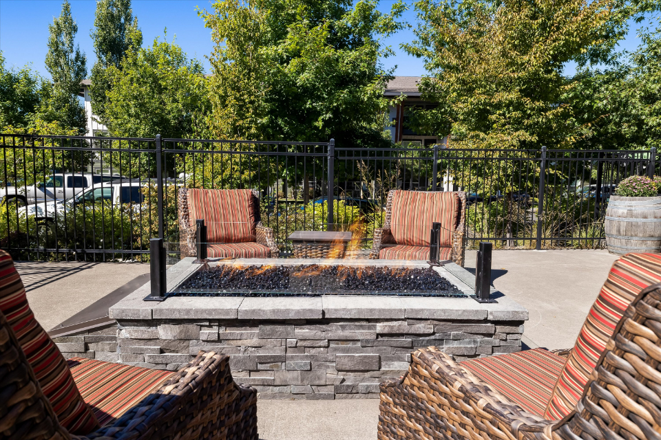 Four cushioned patio chairs surround a rectangular stone fire pit with flames burning, set on a paved outdoor area, with trees, a black metal fence, and cars visible in the background.