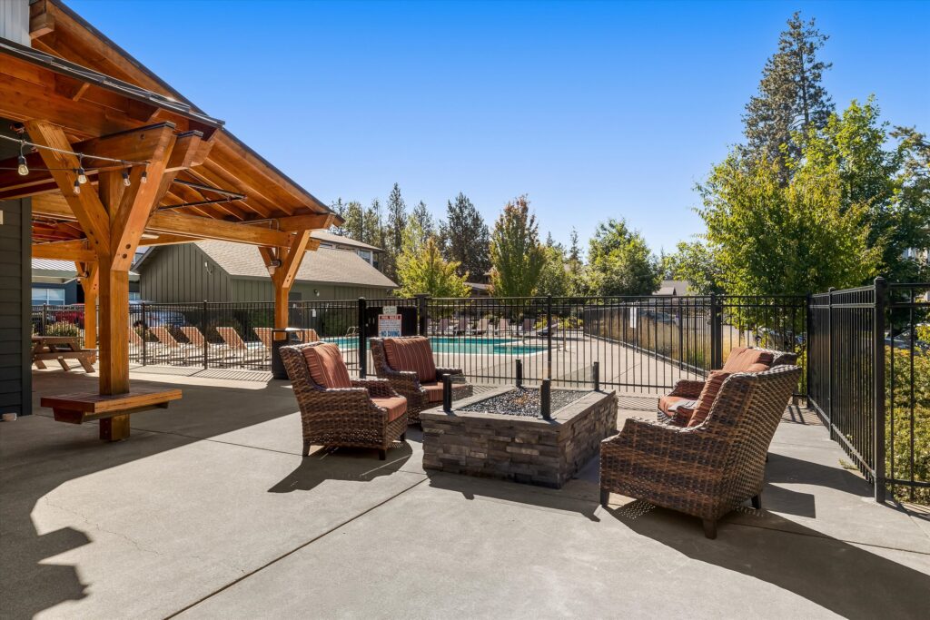 Wicker chairs with cushions surround a fire pit on a patio near a fenced outdoor pool. An open wooden pergola provides shade. Trees and buildings are visible in the background under a clear blue sky.