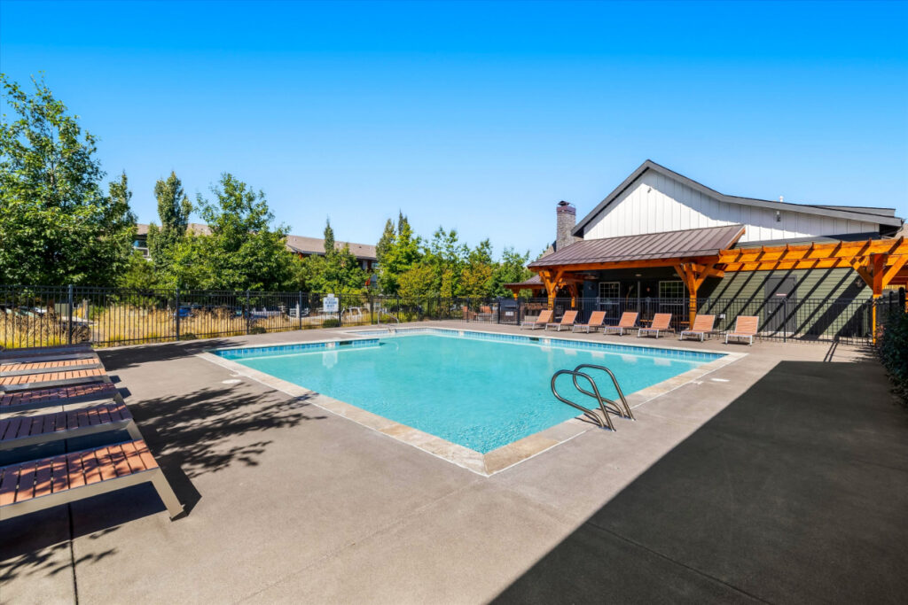 Outdoor swimming pool with clear water, surrounded by lounge chairs and trees, next to a building with a covered patio under a bright blue sky.