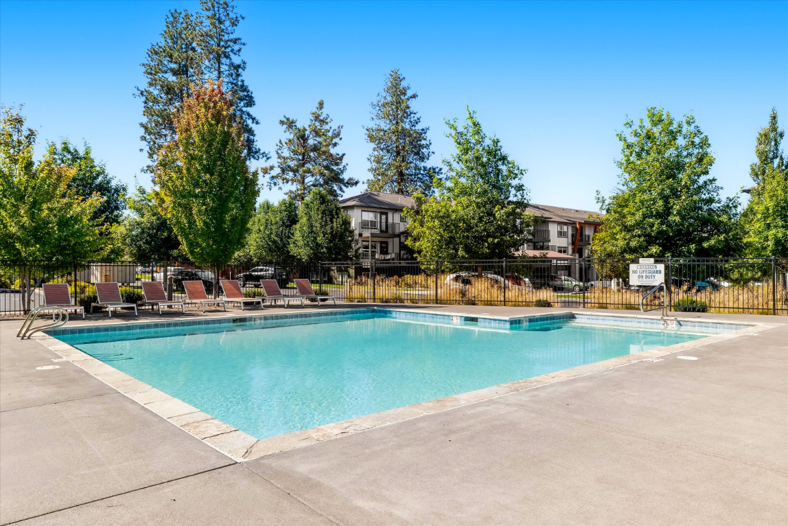 Outdoor swimming pool with clear water, surrounded by lounge chairs, trees, and a fence; apartment buildings and a bright blue sky in the background.