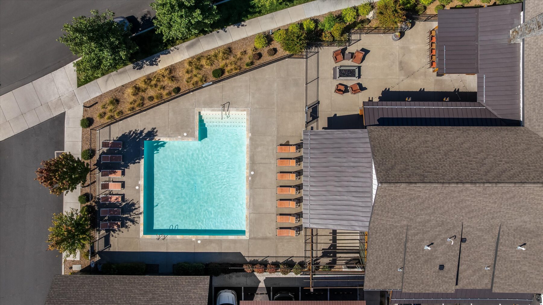 Aerial view of a rectangular outdoor swimming pool with lounge chairs, surrounded by buildings, greenery, and a concrete path.