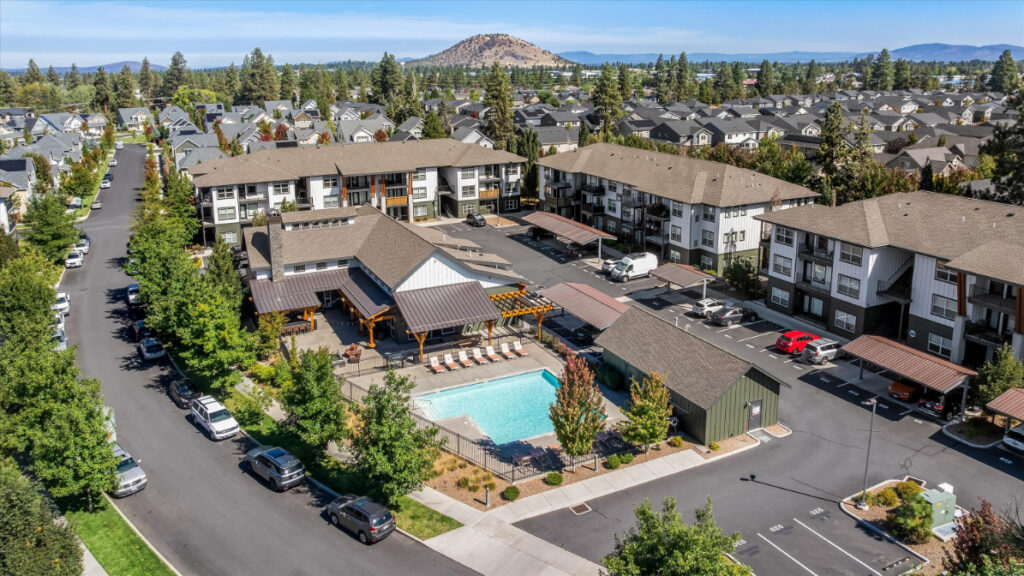 Aerial view of an apartment complex with a central swimming pool, surrounded by trees and parked cars, with a mountain visible in the background under a clear blue sky.