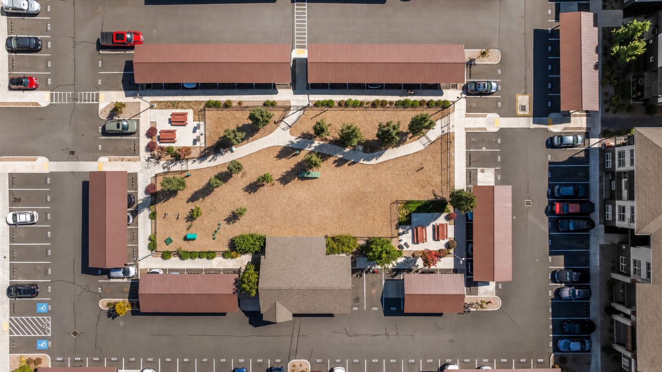 Aerial view of a rectangular park with a brown lawn, trees, and paths, surrounded by parking lots, cars, and buildings. Four covered structures border the park on each side.