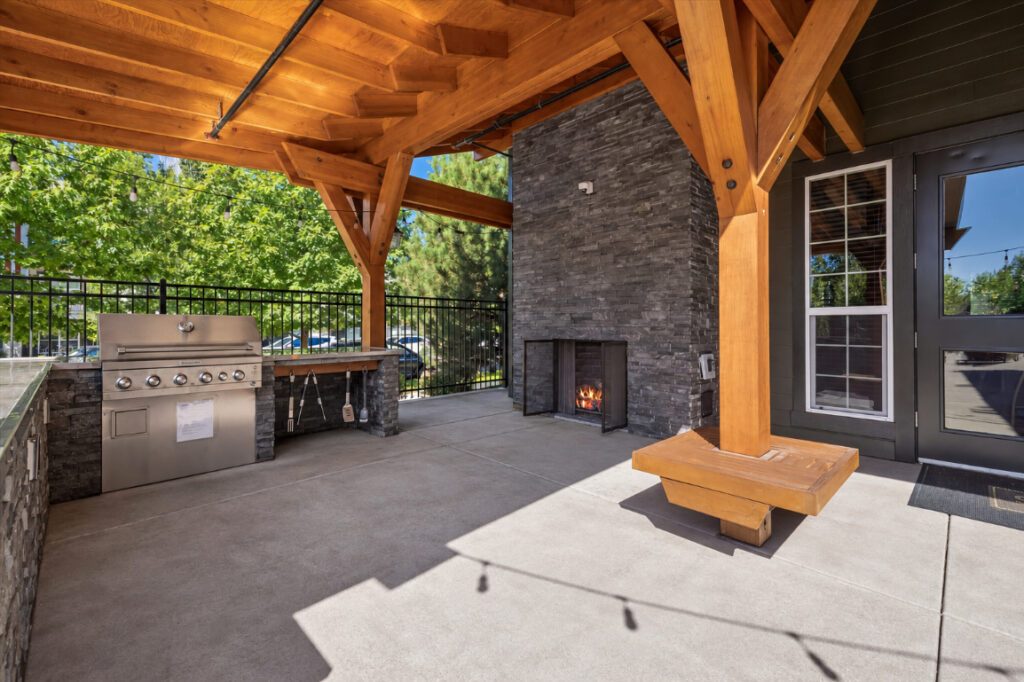 Outdoor patio with a wooden pergola, built-in stainless steel grill, stone fireplace, bench seating, and trees visible in the background behind a black metal fence.