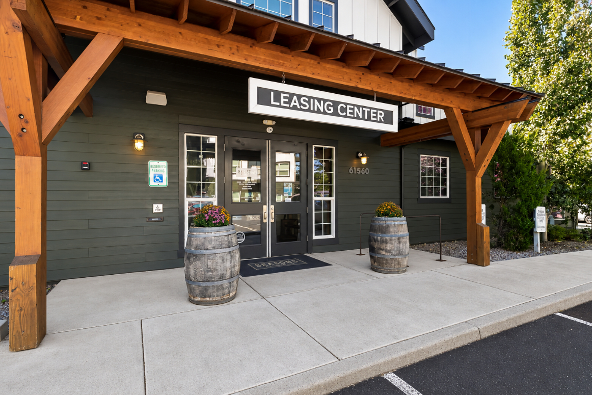 Entrance to a leasing center with a wooden awning, two large planters, a welcome mat, and a sign above the door. Glass doors and windows display the address 61510.